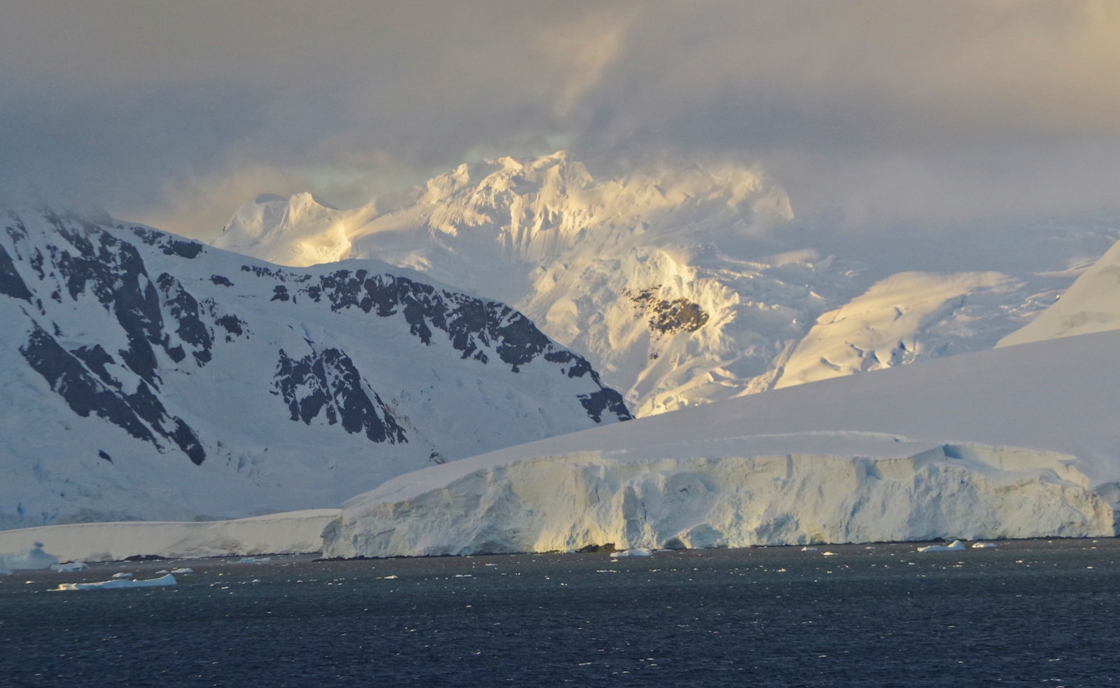 An expedition cruise ship navigating between large blue and white icebergs in calm Antarctic waters at sunset with the Antarctic peninsula