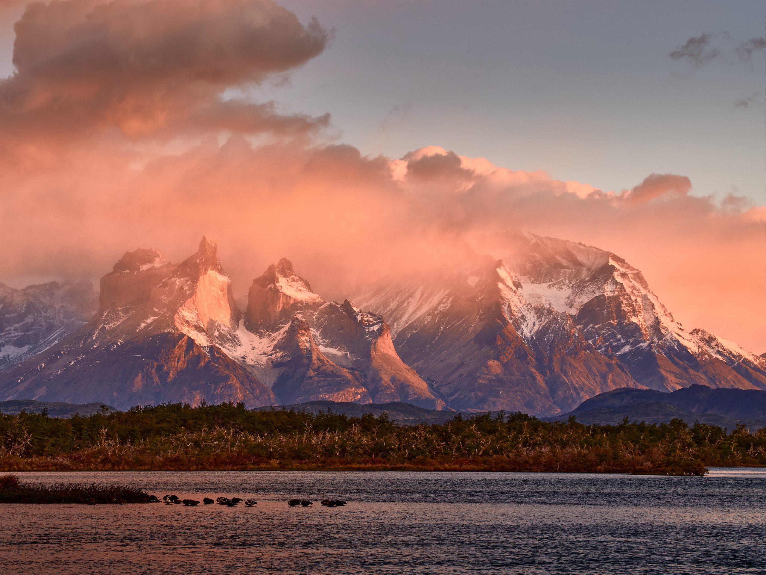 The Torres del Paine massif at sunrise with the granite spires lit in pink and orange light against a clear sky, the dark waters of a glacial lake in the foreground reflecting the peaks.